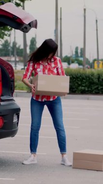 Kadın kurye, kırmızı ekose gömlekli ve kotlu, paketleri boşaltıyor, bagajdan karton kutular çıkarıyor, açık havada. Kargo teslimat servisi..