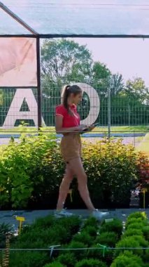 Kadın bahçıvan bitkilerin kalitesini kontrol eder, dijital tablete not alır, bahçe merkezi ya da sera boyunca yürür. Bitkileri büyütmek ve onlarla ilgilenmek. Çiçekçilik, bahçıvanlık, bahçıvanlık, çiçek işi..