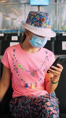 Koruyucu maskeli genç kız cep telefonu kullanıyor. Boş bir havaalanı salonunda oturmuş, uçağa binmeyi bekliyor. Coronavirüs salgını bittikten sonra uçuşlar yeniden başladı. Sınırlar açılıyor. seyahat açık.