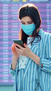 Hava alanında kalkış panosuna karşı duran maskeli kadın yolcu. Uçuş saatlerini cep telefonuyla kontrol ediyor. Coronavirüs salgınından sonra hava yolculuğu açılıyor..