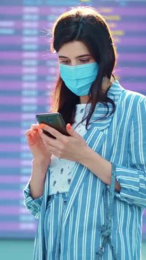 Hava alanında kalkış panosuna karşı duran maskeli kadın yolcu. Uçuş saatlerini cep telefonuyla kontrol ediyor. Coronavirüs salgınından sonra hava yolculuğu açılıyor..