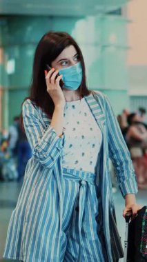 Havaalanında maskeli bir kadın, hava yolcusu kalabalığının arka planında telefonla konuşuyor. Uçağa binmek için bekliyor, bavullarıyla. Coronavirüs salgınından sonra hava yolculuğu açılıyor..