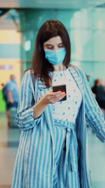 Maskeli kadın yolcu, havaalanında duruyor, bavullarıyla uçağa binmek için bekliyor. Uçuş saatlerini cep telefonuyla kontrol ediyor. Coronavirüs salgınından sonra hava yolculuğu açılıyor..