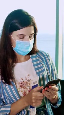 Maskeli kadın portresi, mobil kullanıyor, panoramik pencerenin önünde oturuyor, havaalanında büyük uçak manzaralı. Coronavirüs salgını bittikten sonra uçuşlar yeniden başladı. Sınırlar açılıyor. seyahat açık