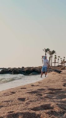 Koruyucu maskeli ve yazlık elbiseli bir adam, deniz kenarında, sıcak güneş ışınlarını, palmiye ağaçlarıyla dolu plajları ve seyahat çantasını seviyor. Covid-19 'un peşinden gidiyorum. kendi kendini izole etmenin sonu .