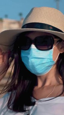 Koruyucu maskeli genç bir kadın, yaz kıyafetleri, güneş gözlükleri ve güneş şapkası, marinadaki bir yatın arka planında. Yat sezonu açıldı, Covid-19 'un peşinden gidiyorum.