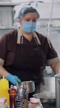 Yemek pişirmek. Kadın şef, koruyucu eldivenler ve maske takmış, mutfakta yemek hazırlıyor, kantinde. Sağlıklı gıda ve güvenlik kavramı. Matarayı yeniden aç