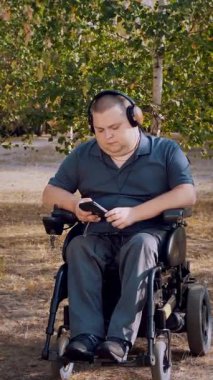 Tekerlekli sandalye. Engelli adam. Genç engelli bir adam, otomatik tekerlekli sandalyede oturuyor ve güneşli bir sonbahar gününde şehir parkında kulaklıkla müzik dinliyor..
