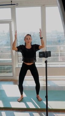 Bayan fitness koçu akıllı telefon kullanarak online egzersiz gösteriyor. Tecrit sırasında zindelik. Evde spor yapan genç bir kadın. Zindelik, sağlık ve sağlıklı yaşam tarzı kavramı