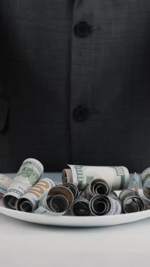 İş adamı ve para, nakit. Bir iş adamı, resmi bir iş elbisesi içinde elinde çatal, bıçak ve çatalla tabakta yüz dolarlık banknotlar yuvarlıyor. Para yiyecek gibi görünüyor..