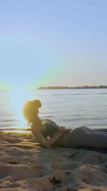 Barda idman. Açık havada uzanıyor. Yoga kumsalı. Atletik genç kadın gün batımında ya da gün doğumunda plajda minder üzerinde egzersiz yapıyor. Spor salonu açık havada. Fitness, bale, spor, yoga