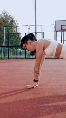 Spor salonu açık havada. Beyaz bluzlu, şortlu, turuncu spor ayakkabılı ve güneş gözlüklü sportif bir kadın, turuncu sahada beyaz işaretli çeşitli egzersizler yapıyor. Yaz günü. Açık hava sporları