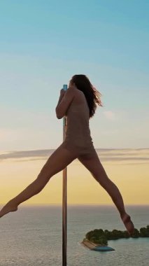 Direk dansı. Silüet. uygun kadın jimnastikçi, bej taytlı, akrobatik egzersizler yapıyor metal döner direkte, gökdelen çatısında, deniz manzaralı arka planda