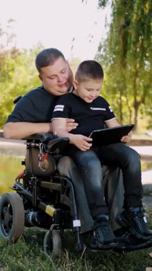 Adam tekerlekli sandalye kullanıcısı. Baba ve oğul. Babalar Günü. Küçük oğluyla birlikte bir adam, bir yaz günü, parkta dizüstü bilgisayarından bir şey izliyor. Eğleniyorum..