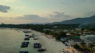 İHA iniyor. Muhteşem bir Tayland plajının üzerinde güneş batıyor, turistler manzaranın tadını çıkarıyor. Turuncu ve altın rengi gökyüzü okyanusa sıcak bir ışık saçıyor. Yaz tatili için mükemmel bir yer.. 