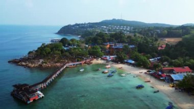 Turkuvaz suları ve yemyeşil yağmur ormanlarıyla çevrili, yüzen bir restorana giden uzun ahşap bir iskelesi olan tropik bir adanın havadan görünüşü. panorama genel görünüm dronu 