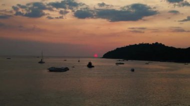 Güneş sakin okyanusun üzerinde batıyor, teknelere sıcak bir parıltı saçıyor ve uzaktaki bir ada, gökyüzünü turuncu ve pembe tonlarında boyuyor. panorama genel görünüm dronu 