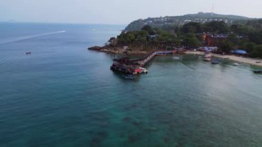 Turkuvaz suları ve yemyeşil yağmur ormanlarıyla çevrili, yüzen bir restorana giden uzun ahşap bir iskelesi olan tropik bir adanın havadan görünüşü. geniş yörünge görünümü dronu 