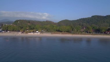 Panorama insansız hava aracı. Turistler gün batımında kumsalda yürürken arka planda yemyeşil bir tropikal orman görüyorlar.