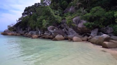 İHA yükseliyor. Turkuaz okyanus suyu, tropik bir adada yemyeşil bir ormanı olan kayalık bir sahille buluşuyor.