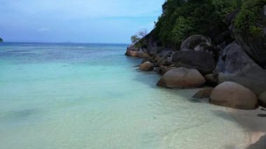 Üstten kumandalı hava aracı. Turkuaz okyanus suyu, tropik bir adada yemyeşil bir ormanı olan kayalık bir sahille buluşuyor.
