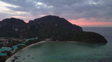 İniş yapan insansız hava aracı, tropikal bir adanın dramatik gökyüzü ile gün batımında tatil beldelerini ve tekneleri gösteriyor.