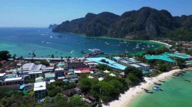 Turkuaz su, tropikal bir adanın kıyılarında nazikçe dolaşırken, arka planda deniz manzarasını noktalayan kayıklar ve yemyeşil tepeler yükseliyor. Sağ drona döndür 