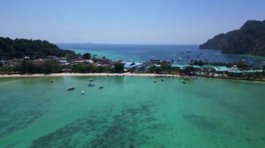 Turkuaz su, tropikal bir adanın kıyılarında nazikçe dolaşırken, arka planda deniz manzarasını noktalayan kayıklar ve yemyeşil tepeler yükseliyor. panorama yörünge dronu 