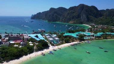 Turkuaz su, tropikal bir adanın kıyılarında nazikçe dolaşırken, arka planda deniz manzarasını noktalayan kayıklar ve yemyeşil tepeler yükseliyor. panorama genel görünüm dronu 