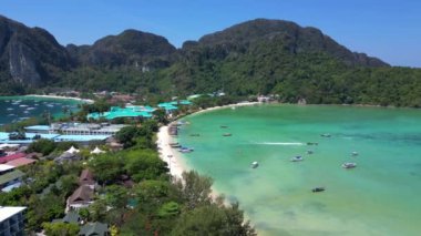 Turkuaz su, tropikal bir adanın kıyılarında nazikçe dolaşırken, arka planda deniz manzarasını noktalayan kayıklar ve yemyeşil tepeler yükseliyor. uçuş üstü insansız hava aracı 