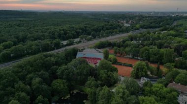 Kırmızı kil kortları olan, yemyeşil bir orman ve küçük bir göl ile çevrili bir tenis stadyumunun havadan görünüşü. uçuş üstü insansız hava aracı 