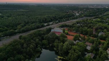 Kırmızı kil kortları olan, yemyeşil bir orman ve küçük bir göl ile çevrili bir tenis stadyumunun havadan görünüşü. İHA alçalıyor 