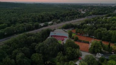 Kırmızı kil kortları olan, yemyeşil bir orman ve küçük bir göl ile çevrili bir tenis stadyumunun havadan görünüşü. Ters İHA uçuşu 