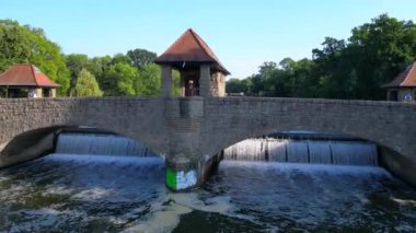 Palmengartenwehr Elster Weir, parlak bir yaz gününde yemyeşil ağaçlarla çevrili bir köprünün altından akan nehrin havadan görünüşü. uçuş üstü insansız hava aracı 