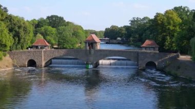Palmengartenwehr Elster Weir, parlak bir yaz gününde yemyeşil ağaçlarla çevrili bir köprünün altından akan nehrin havadan görünüşü. uçuş üstü insansız hava aracı 