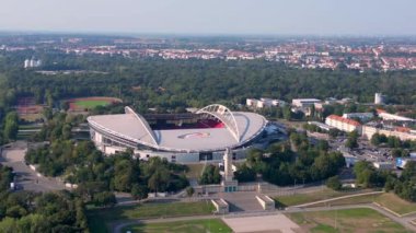 Güneşli bir yaz gününde Rb Leipzig 'e ev sahipliği yapan bir arenanın hava manzarası yemyeşil çevreyi ve şehir manzarasını gözler önüne seriyor. Ters İHA uçuşu 