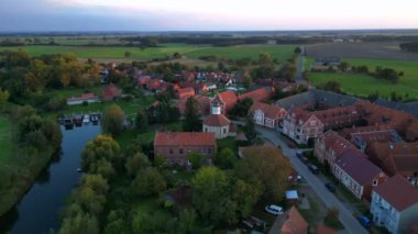 Altın saat ışığıyla aydınlatılan küçük bir köyün havadan görünüşü, etrafındaki tarlaların üzerine uzun gölgeler düşürüyor. panorama yörünge dronu 