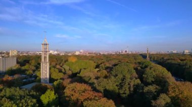 Renkli sonbahar yapraklarıyla çevrili ikonik bir dönüm noktası, açık mavi gökyüzünün altında Berlin şehri manzarası. Yukarıdan insansız hava aracı görüntüleri 