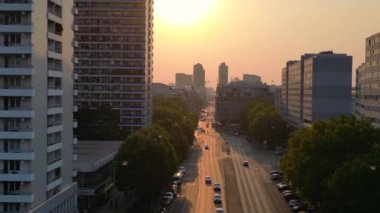 Gün batımında bir bulvarda giden arabalar, ağaçlarla ve apartmanlarla dolu bir şehir manzarasında. İticileri ileri çalıştır 