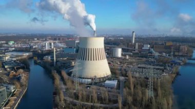 Güneşli bir günde, arka planda bir nehir ve şehir bulunan bir termik santralin dumanı yayan hava görüntüsü. İHA alçalıyor 