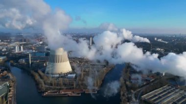 Güneşli bir günde, arka planda bir nehir ve şehir bulunan bir termik santralin dumanı yayan hava görüntüsü. İHA alçalıyor 