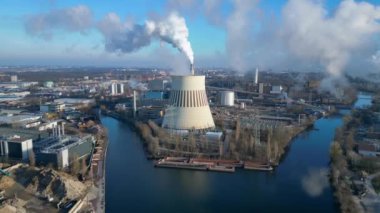 Güneşli bir günde, arka planda bir nehir ve şehir bulunan bir termik santralin dumanı yayan hava görüntüsü. İticileri ileri çalıştır 