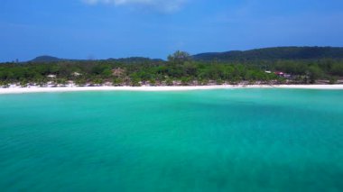Beyaz kumu yıkayan turkuaz suyla Sok San Sahili 'nin havadan görünüşü. Palmiye ağaçları ve tropikal bitki örtüsüyle çevrili. İticileri ileri çalıştır 