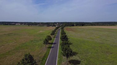 Ağaçlar ve tarlalarla dolu kırsal bir araziden geçen boş bir asfalt yolun havadan görünüşü. Ters İHA uçuşu 