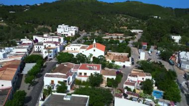 İspanya 'nın İbiza kentindeki Sant Joan de Labritja' da, güneşli bir yaz gününde geleneksel beyaz kilise, evler, yollar, dağlar ve yemyeşil alanlar yer alıyor. panorama yörünge dronu 