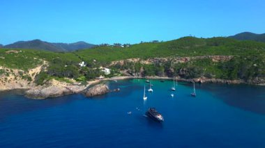 İspanya 'nın İbiza kentindeki yemyeşil bir körfezin turkuaz sularında demirlenmiş yelkenli ve yatların çarpıcı hava manzarası. İHA yükseliyor 