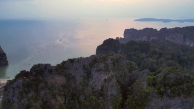 Andaman Denizi 'ni noktalayan geleneksel uzun kuyruklu tekneleriyle gün doğumunda yükselen kireçtaşı kayalıklarıyla çevrili Railay Sahili, Krabi, Tayland. panorama genel görünüm dronu 