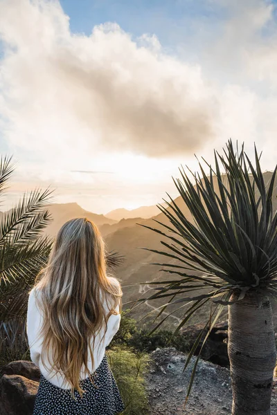Rear view of blonde woman in a sunset sky and sun through the clouds. Tranquil landscape and horizon over the mountains