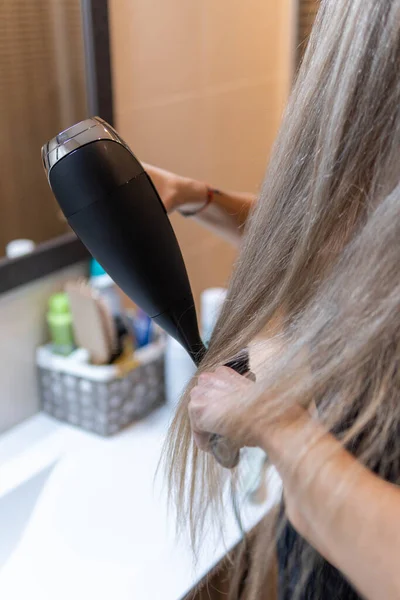 Unrecognizable blonde woman with wet hair combing her hair with dryer in modern bathroom in front mirror