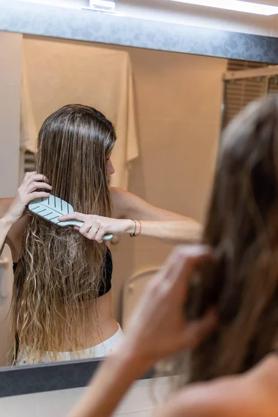 Blonde woman with wet hair combing her hair in modern bathroom in front of mirror
